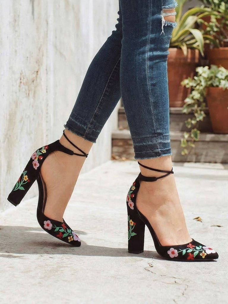 Black block-heeled shoes with floral embroidery worn with blue jeans on a light background.