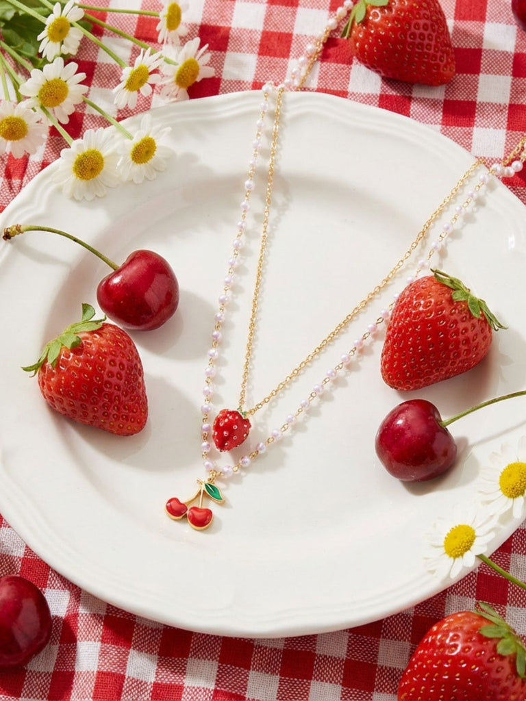 Necklace with strawberry and cherry details on a white plate with strawberries and cherries, on a red and white checkered tablecloth.