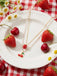 Necklace with strawberry and cherry details on a white plate with strawberries and cherries, on a red and white checkered tablecloth.