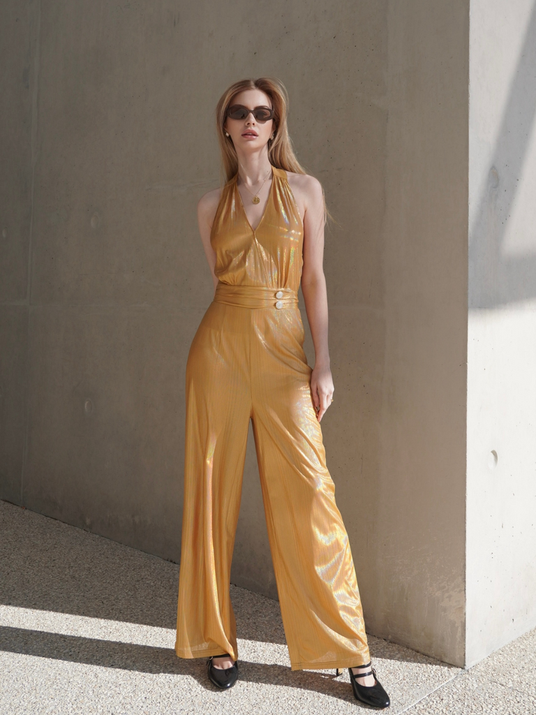 A woman wearing a gold V-neck halter jumpsuit with wide-leg pants, paired with sunglasses and black shoes.