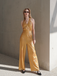 A woman wearing a gold V-neck halter jumpsuit with wide-leg pants, paired with sunglasses and black shoes.