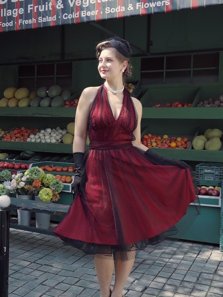 A burgundy 1950s style swing dress with a black polka dot mesh overlay, featuring a halter neck and sleeveless design, shown with a fashion illustration.