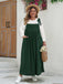 A plus-size woman wearing a forest green sleeveless pinafore maxi dress over a white long-sleeve top, styled with a straw hat and bag.