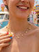 A smiling woman wearing the gold ocean charm necklace outdoors at a carnival or beach setting.