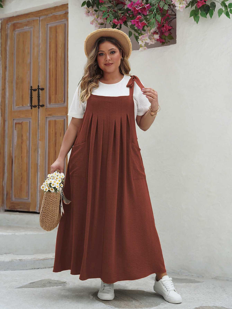 Front view of the pinafore-style maxi dress in rust brown featuring shoulder ties and a pleated skirt.